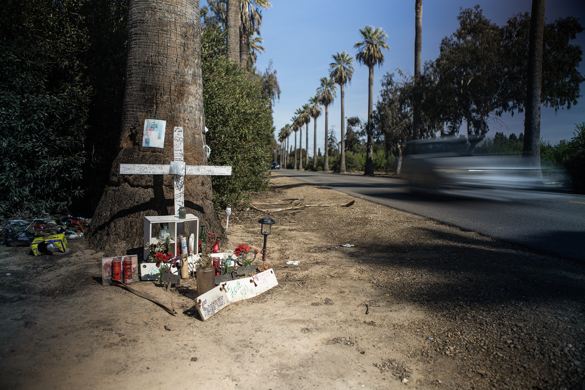 A roadside memorial at the base of a palm tree along a rural highway. A white wooden cross with handwritten messages is placed against the tree, surrounded by candles, flowers, photos, and other personal items. A car drives past in the background, showing how traffic continues near the site of the crash. The image documents a space where people are remembering someone who died.